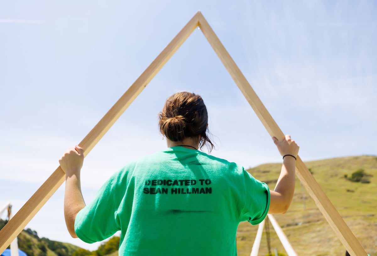 A student wears a green T-shirt that says "Dedicated to Sean Hillman" on the back as they put two wooden beams together.