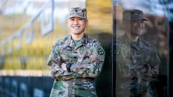 A man in military camoflage smiles in front of a building window.