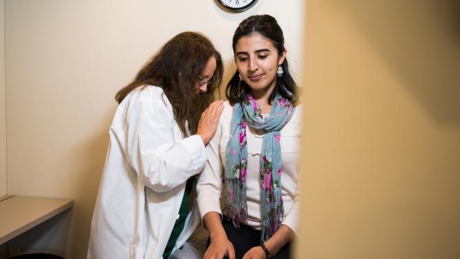 Student seated while a physician listens to their back with a stethoscope.